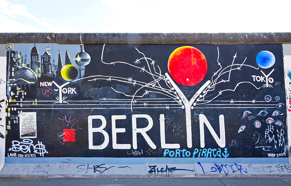 Street art mural on the Berlin Wall depicting connections between New York, Berlin, and Tokyo, with barbed wire and abstract imagery.